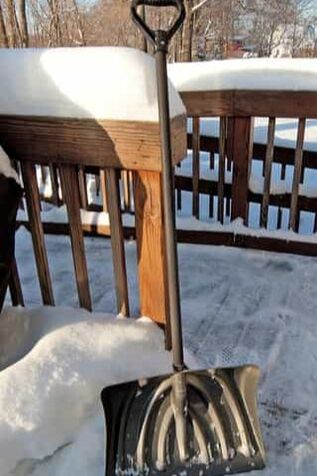 A metalic snow shovel on a wooden deck. there is about a foot of snow on the yard and on the deck.