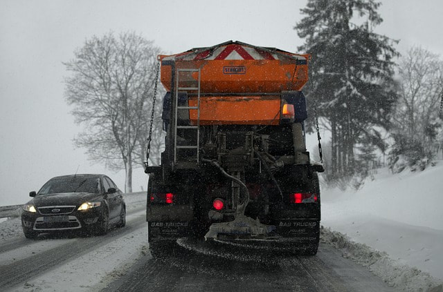 A yellow snow plow is removing snow from someones driveway in Syracuse. There appears to be 2-3 of snow on the ground.