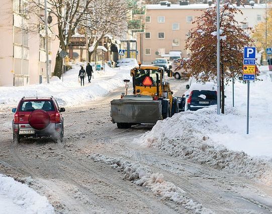 A large orange plow plowing a residential road in downtown Syracuse. There is about two feet of snow on the ground and the plow is clearing the road for several cars as well as dropping salt.