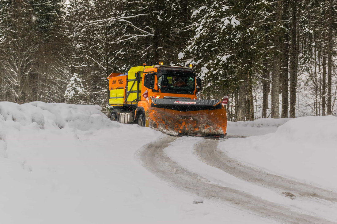 An orange snow plow salting a road while snow falls around it. The plow is out in a rural area and a black Chevrolet sedan is driving by. 