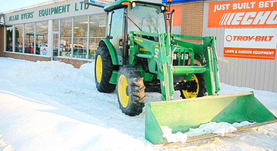 A green plow clears snow from a store front in a commercial area. 
